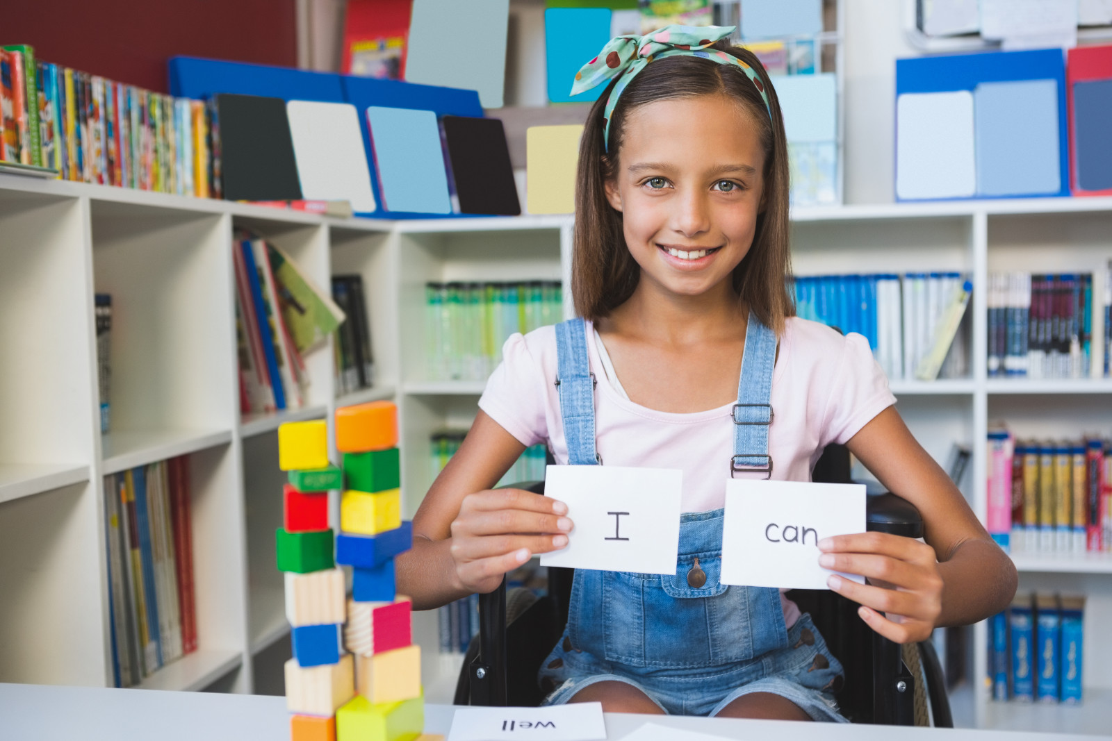 a Hispanic girl on a wheelchair holding up two cards saying I and Can